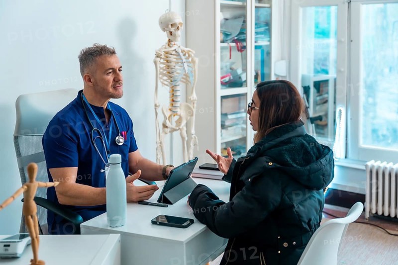 Doctor in blue scrubs and stethoscope consulting a female patient regarding her health concerns, discussing treatment options in a modern medical office setting with a skeleton model
