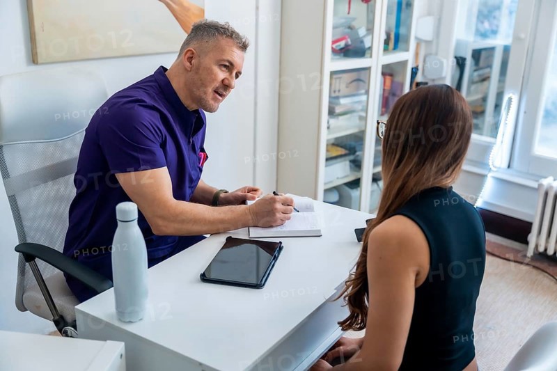 Physiotherapist in purple scrubs discussing treatment plan with a female patient, recording notes in a professional office setting for rehabilitation and care