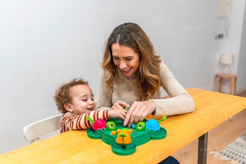 Mother and young son enjoying a playful moment, laughing and interacting while sitting at a wooden table and playing a colorful board game, sharing happiness and bonding