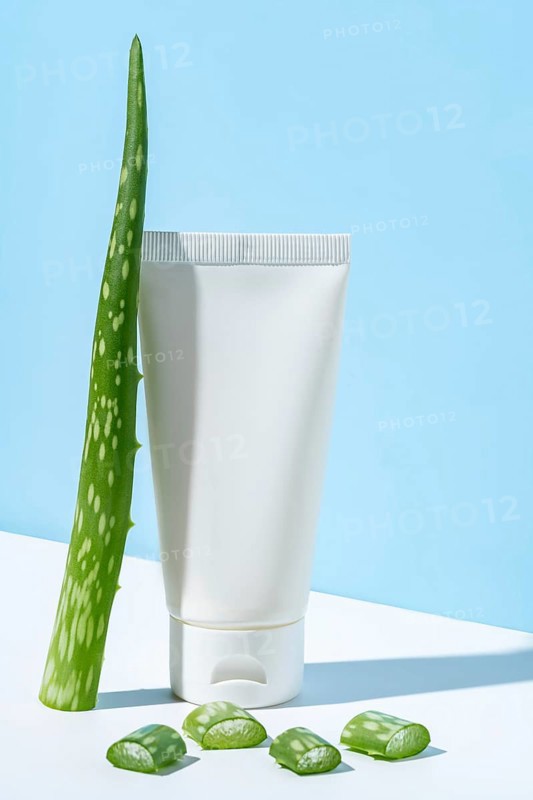 This image portrays a simple and clean representation of beauty and skincare featuring a white tube of aloe vera cream accompanied by a fresh aloe vera leaf and sliced pieces, staged against a soft light blue backdrop