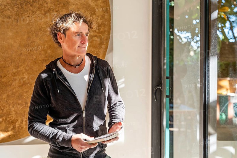 A relaxed man stands indoors holding a book, bathed in natural sunlight near a window Wearing a black hoodie and white t-shirt, he exudes a casual, contemplative vibe at home