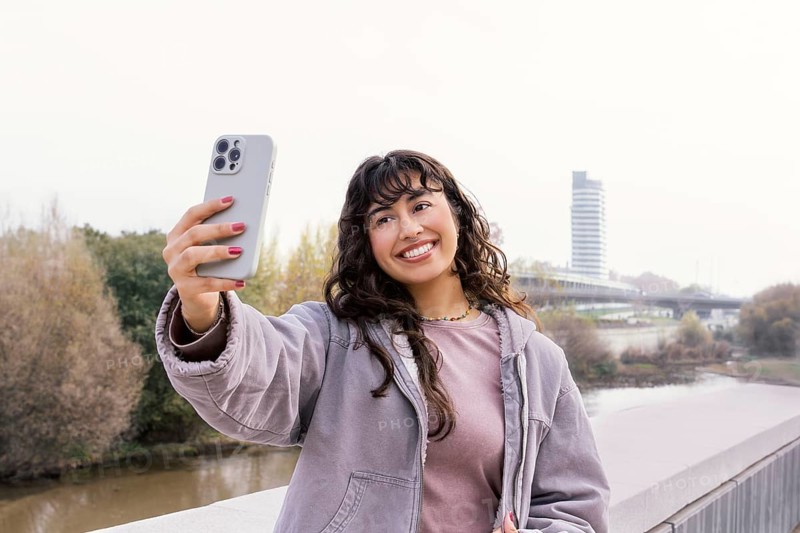 A young Latin woman takes a selfie outdoors, capturing joyful moments The scene is set amidst a natural background, showcasing leisure and happiness