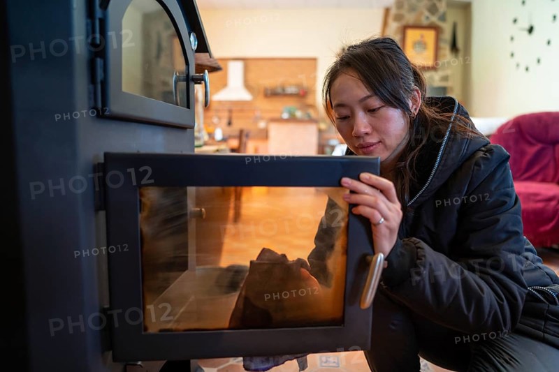 An Asian female looks down while meticulously cleaning the glass door of a wood burning stove in a cozy, well-lit room. Her focused expression and the warm ambiance highlight a moment of comfort and warmth