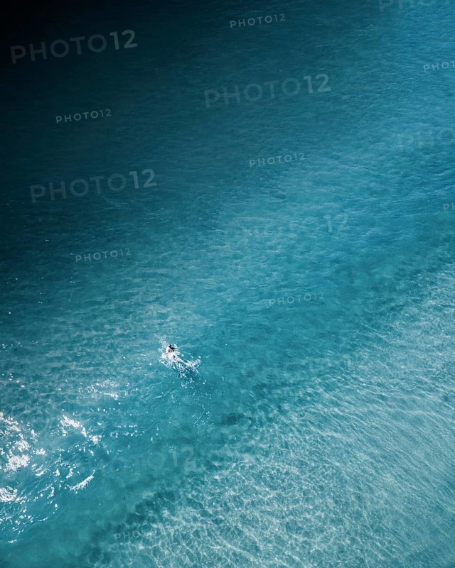 An aerial shot of a lone surfer paddling through calm turquoise waters along the Portuguese coast. The serene and minimalist composition highlights the beauty of surfing in nature