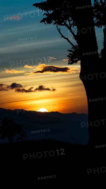 Captivating scene of the sun setting behind the silhouetted mountains, viewed from a vantage point overlooking San Jose, Costa Rica. The sky is painted in vibrant shades of orange and blue, with scattered clouds illuminated by the sunset