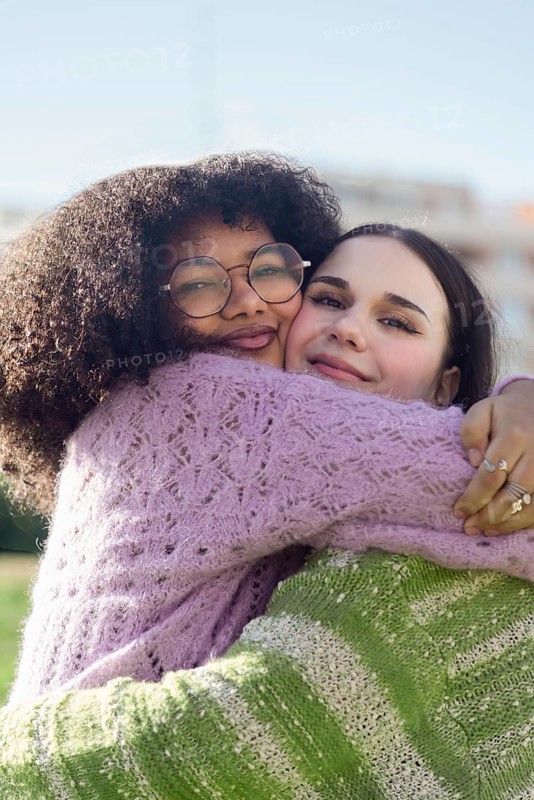 Two friends share a heartfelt hug, beaming with joy in a sunny outdoor setting. Both wear cozy knitted sweaters, radiating a sense of warmth and happiness
