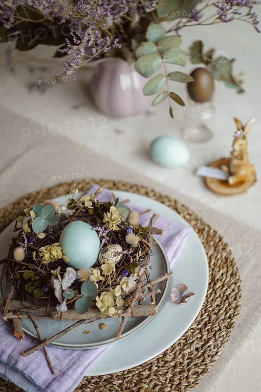 Top view of an Easter table setting featuring a plate with a rustic nest of twigs and naturally dyed eggs, surrounded by floral decorations and subtle tableware accents under soft, natural lighting