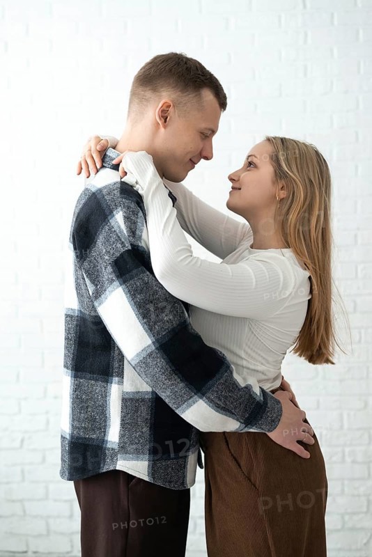 A loving couple shares a warm embrace in a brightly lit room, conveying a sense of happiness and intimacy. Both are smiling gently, clearly enjoying the moment with each other