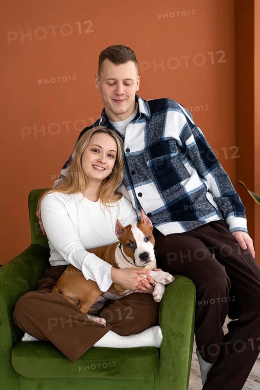 A young Caucasian man and woman smiling, seated comfortably in an armchair with their white and brown dog, in a well-lit room with orange walls