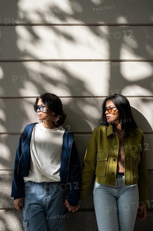 A lesbian couple stands against a wall, holding hands and enjoying the dappled sunlight. Both wear sunglasses and casual jackets, basking in the warmth and moment