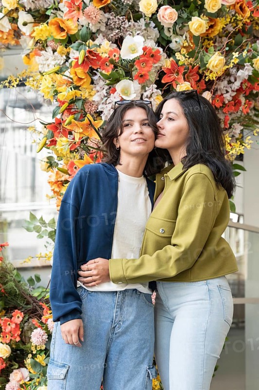 A lesbian couple shares a tender moment in front of a vibrant floral backdrop. Their closeness and happiness are highlighted by the bright, colorful flowers surrounding them