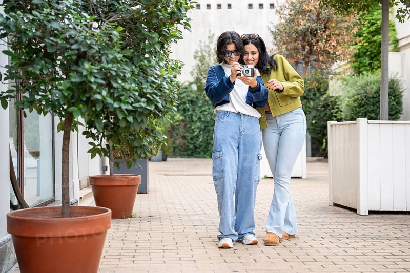 A joyful lesbian couple takes photos in a garden setting. They smile and explore the outdoors, creating cherished memories together with a camera in hand
