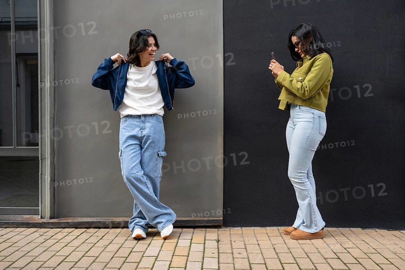 A lesbian couple captures a candid moment outdoors. Both appear happy and relaxed in casual attire against a two-tone wall, sharing a joyful connection