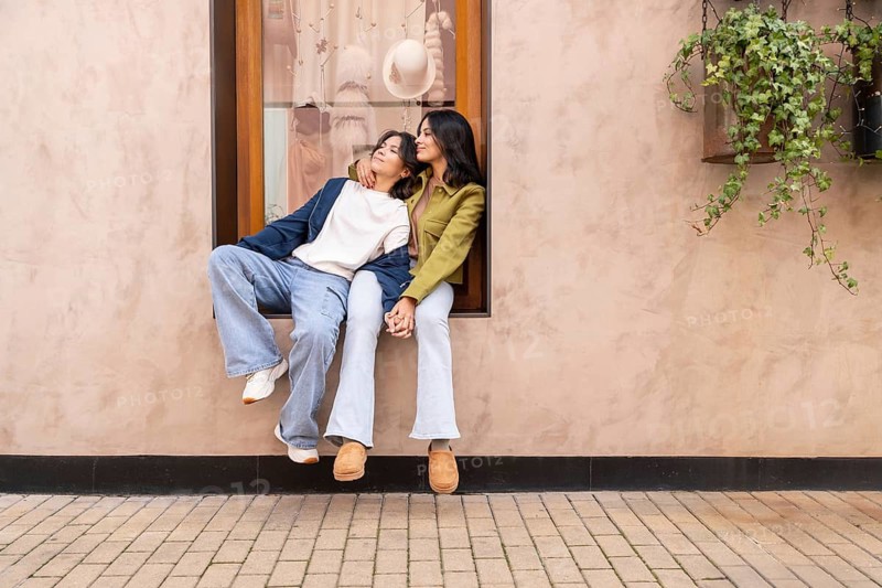 A lesbian couple sits affectionately on a windowsill, holding hands and enjoying a heartfelt moment. The background features a blush-toned wall and a hanging plant