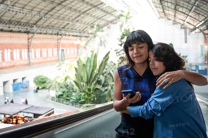 A lesbian couple shares an intimate embrace at Atocha Station in Madrid. One partner hugs the other lovingly while they look at a phone, surrounded by lush greenery and historic architecture