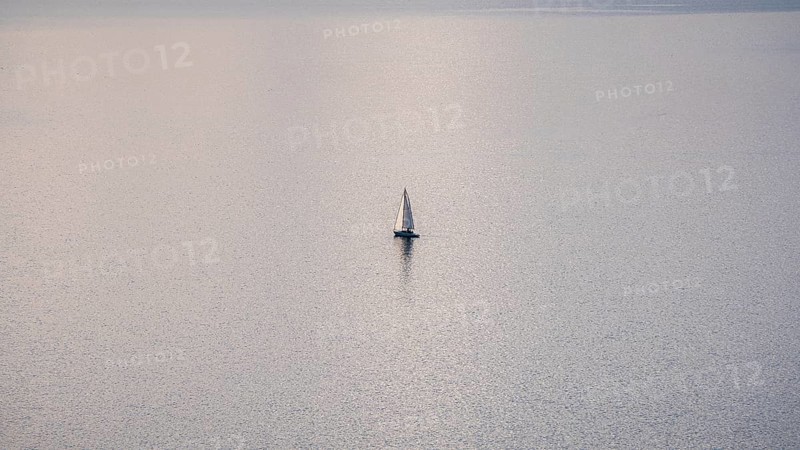 A solitary sailboat on the tranquil waters of Lake Geneva during a sunny winter day, with the snowy Alps looming in the distance, captured from the scenic Lavaux vineyards