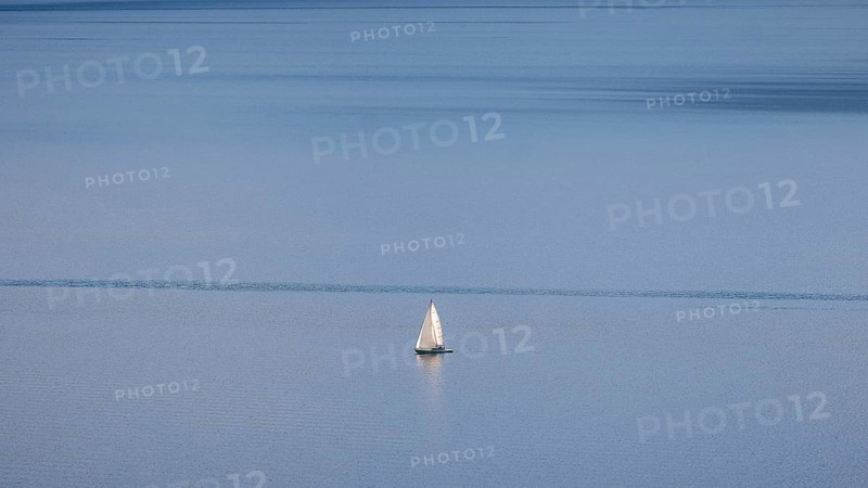 A lone sailboat glides over Lake Geneva on a clear winter's day, framed by the vineyards of Lavaux and distant Alps