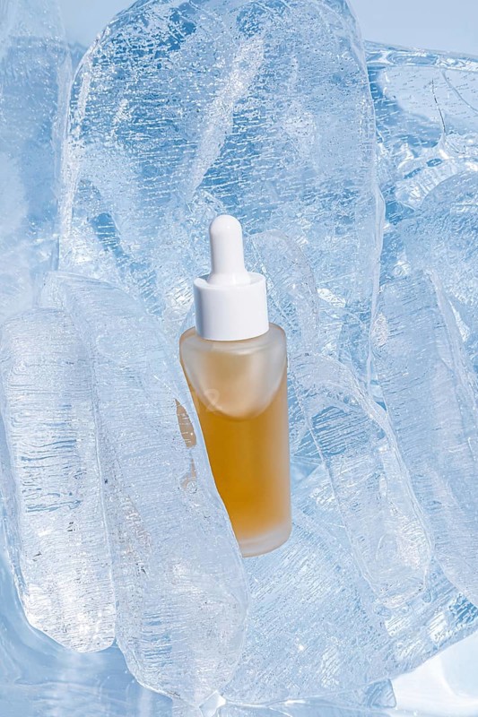 A clear serum bottle with a dropper, encapsulated by shimmering translucent ice cubes, stands out with its golden yellow content