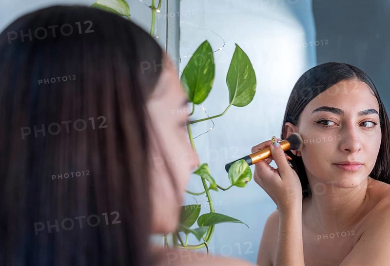 A woman is applying makeup with a brush while looking into a mirror. Green plants add a natural touch to the serene setting, creating a calming atmosphere
