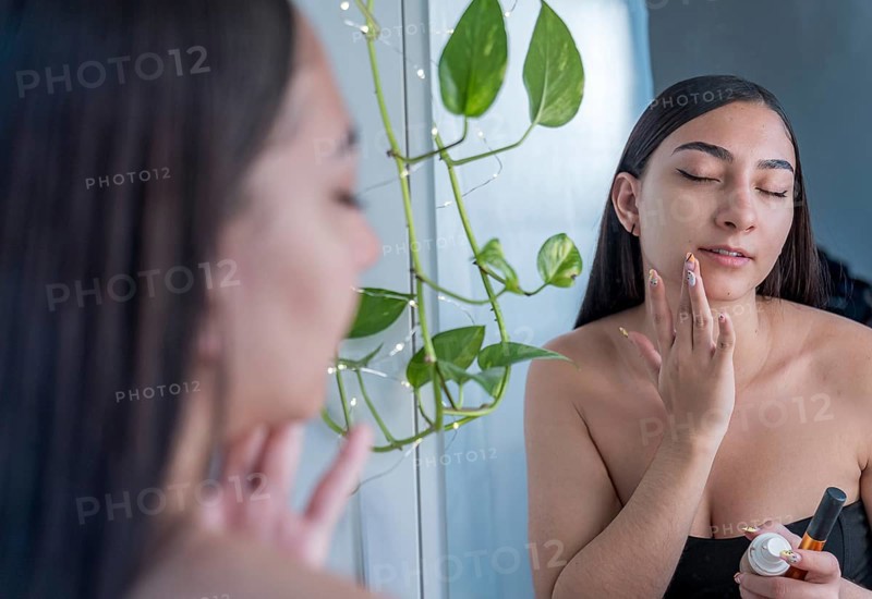 A woman applies skincare cream to her face in front of a mirror. She is focused on self care, with a relaxing atmosphere and natural lighting enhancing her beauty routine