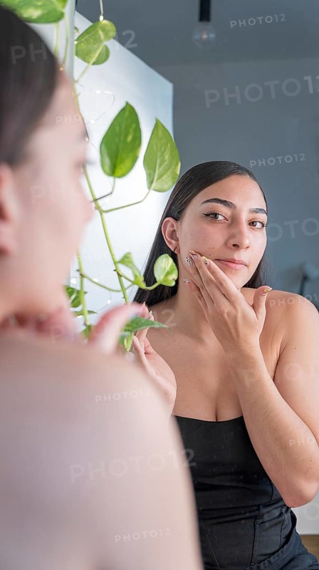 A woman is applying skincare cream while looking in the mirror. Surrounded by indoor plants, she is focused on her self care routine in a peaceful home environment