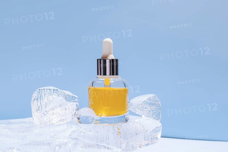 A serum bottle with a dropper, filled with yellow fluid, delicately placed amongst transparent ice chunks against a uniform blue backdrop, highlighting the freshness or cooling aspect of the product