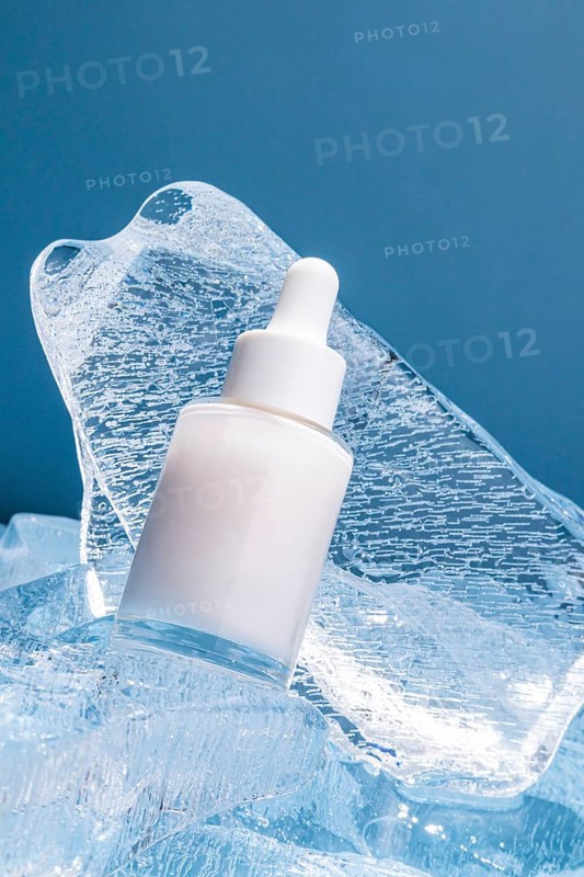 A serum bottle with a dropper rests on textured melting ice cubes against a blue background, conveying a sense of cool freshness