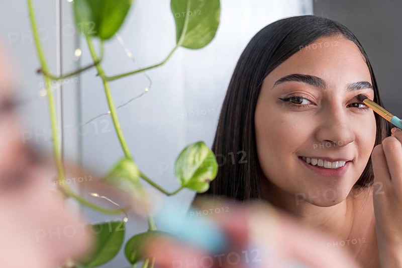 A woman is seen smiling while applying eye makeup in front of a mirror for the makeup session. A green plant is visible, adding a touch of nature to the scene and enhancing the composition