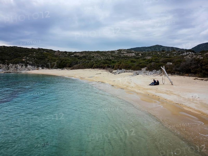 A peaceful aerial view of Kalamitsi wild beach in Greece. A couple sits on the golden sand by turquoise waters, surrounded by lush greenery and a serene natural landscape under a cloudy sky
