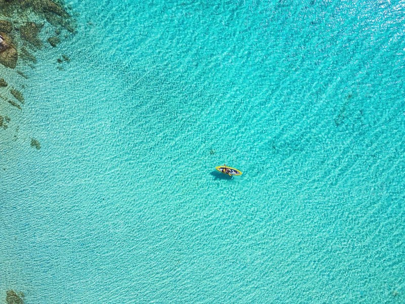 A stunning aerial view of Diaporos Island in Greece, showcasing crystal-clear turquoise waters with a lone kayak drifting peacefully. A paradise of wild beaches and untouched nature