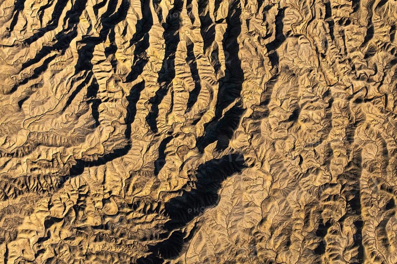 Aerial photograph showcasing intricate golden desert textures at Caineville Mesa, Utah. The landscape features unique erosion patterns under sunlight, creating striking shadows