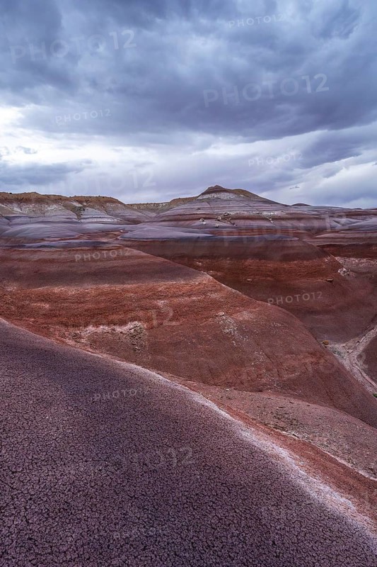 The vibrant, layered Hanksville Bentonites in Utah's desert landscape stretch under dramatic, cloudy skies. The rich colors and textures create an awe-inspiring natural scene