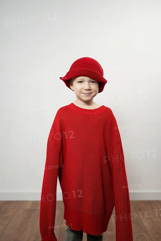 A child wears an oversized red sweater and matching hat, smiling playfully. The neutral background highlights the vibrant clothing, emphasizing a whimsical and cozy theme