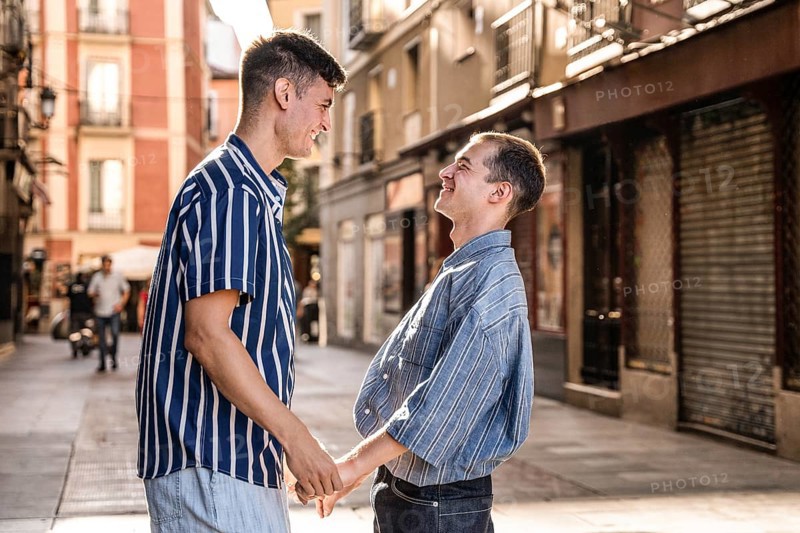 A joyful gay couple shares a moment holding hands on the lively streets of Madrid. The warm light enhances the colorful city backdrop, celebrating love and pride
