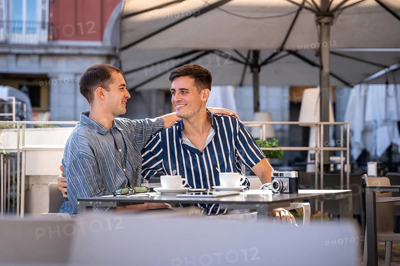 A gay couple shares a joyful moment at a charming outdoor cafe on the streets of Madrid. They are seated at a table, enjoying each other's company in a sunny ambiance