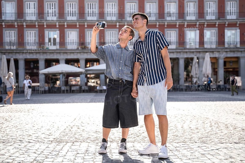 A gay couple enjoys a sunny day in Madrid, capturing memories on the bustling streets while holding hands. The scene reflects love, pride, and togetherness