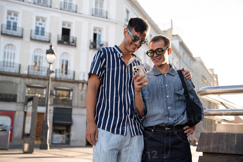 A gay couple enjoys a sunny day, exploring the lively streets of Madrid together. Smiling and looking at a phone, they embody love and connection in an urban setting