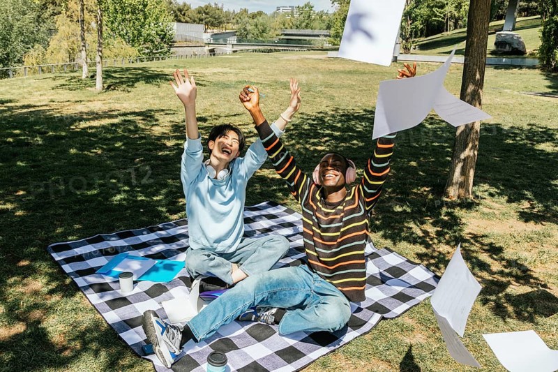 Two university students celebrate joyfully in a sunny park. Sitting on a picnic blanket, they toss papers into the air, enjoying a moment of triumph and relaxation