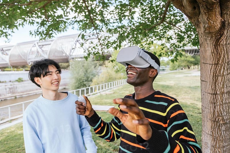Two friends enjoy a VR experience outdoors. One wears a VR headset, immersed in a virtual world, while the other smiles, holding the controller. Trees and a bridge create a serene backdrop