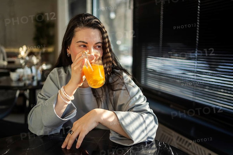 A mid-aged woman sips an orange drink in a brightly lit cafe, her expression serene and content as sunlight streams through the blinds