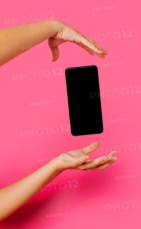 Crop anonymous person in casual clothes showing modern smartphone with empty screen while floating between hands on pink background