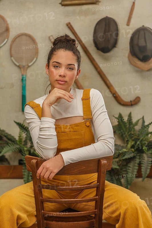 A young woman leans on a vintage wooden chair, dressed in a mustard yellow jumpsuit with a playful air, surrounded by rustic decor and sporting equipment on the walls