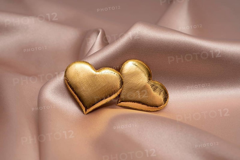 Two golden heart-shaped objects rest gently on a smooth, silky fabric, symbolizing love and commitment, perfect for St. Valentine's Day and wedding themes