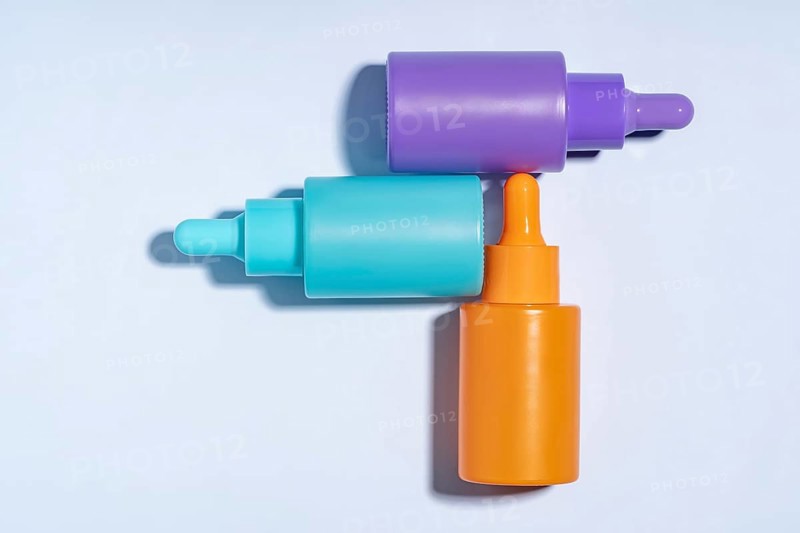 Top view of three vibrant serum bottles in purple, turquoise, and orange colors, designed with droppers, displayed on a clean, light blue background