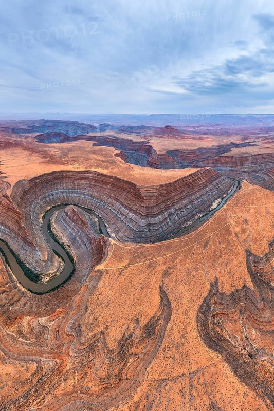 Aerial view of San Juan canyon with a winding river cutting through the arid desert landscape. The layered rock formations display earthy tones under a wide, partly cloudy sky in Utah, USA