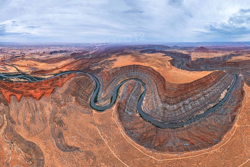 A stunning aerial view captures a meandering river cutting through the rugged San Juan canyon. Layers of red and brown rock form the dramatic landscape under a cloudy sky in Utah, USA