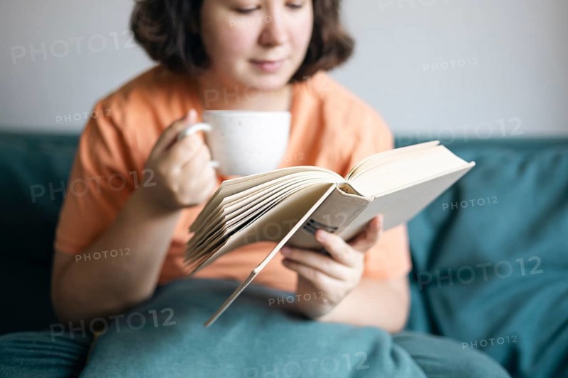 Cropped view of a woman sipping tea and reading a book, comfortably seated on a sofa