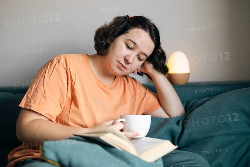Young woman enjoys a quiet moment at home, lounging on a couch with a book and a cup of coffee. She appears thoughtful and relaxed in a cozy domestic setting