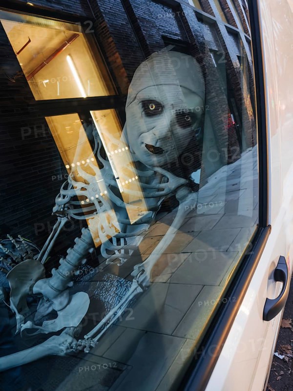 A haunting skeleton figure peers through a city window, its eerie reflection captured on the glass. The scene blends urban architecture with Halloween-style themes