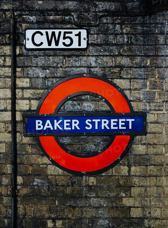 A vibrant image of the Baker Street station sign on London's Underground, set against a textured brick wall. The sign's classic design highlights the station's historical significance